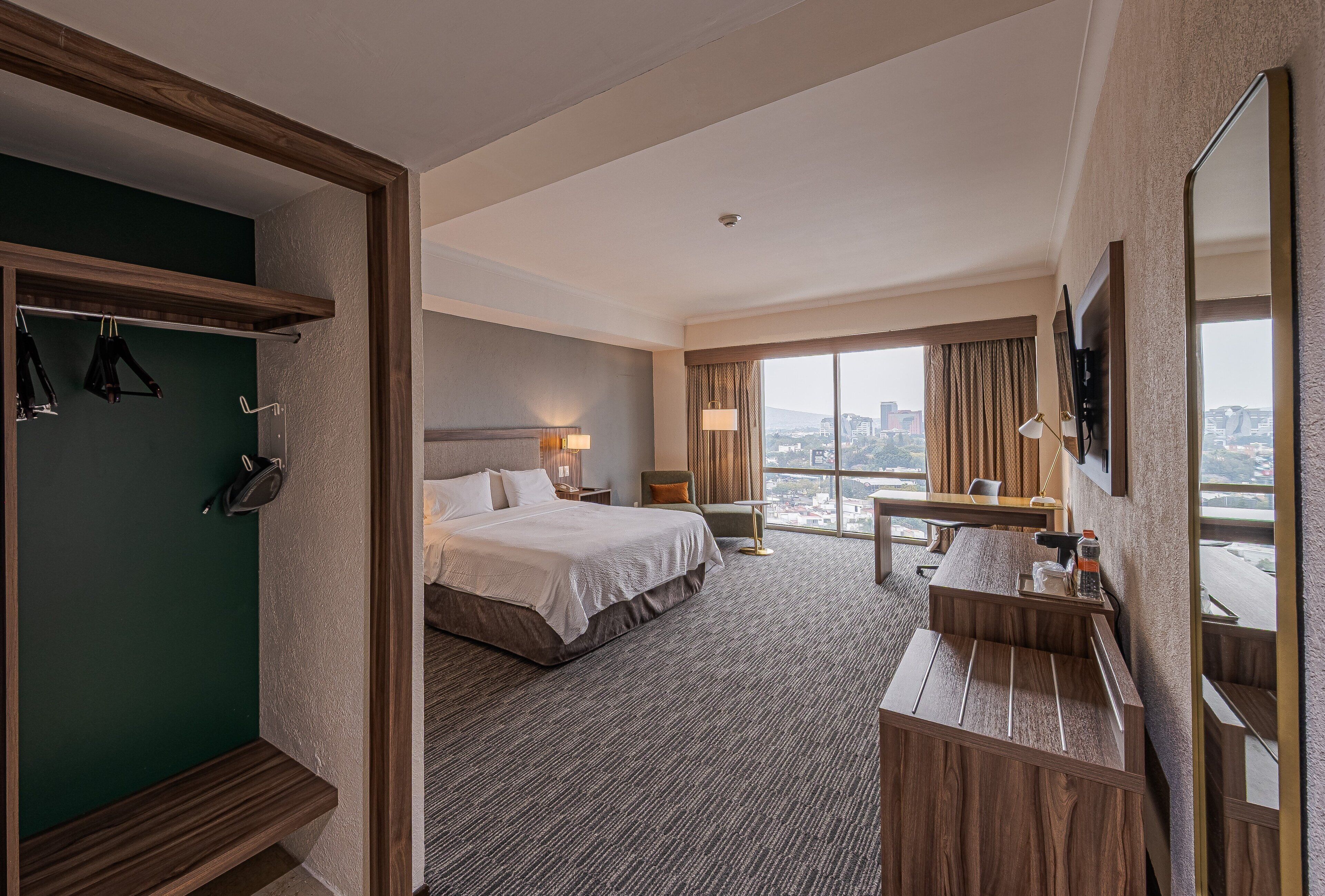 Habitación de hotel con cama, escritorio, silla, televisor, armario y ventana grande con vistas a la ciudad.