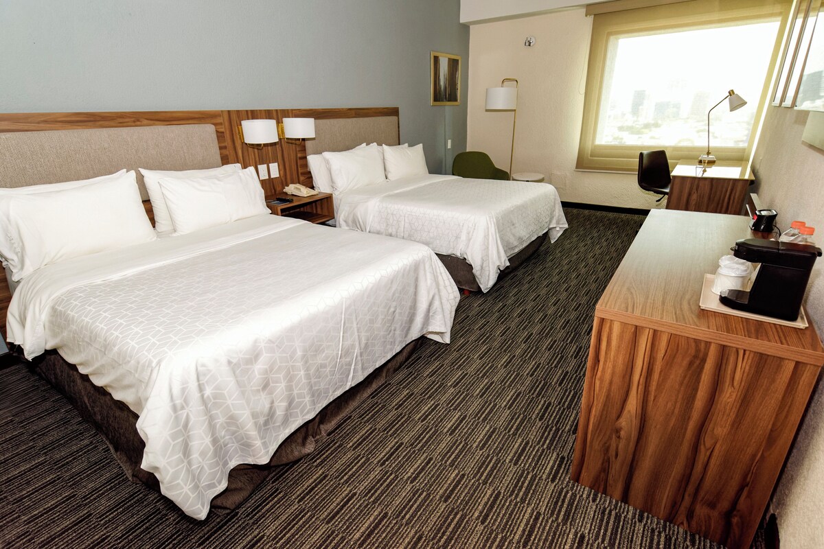 A hotel room with two beds, a desk, a chair, a lamp, and a window.