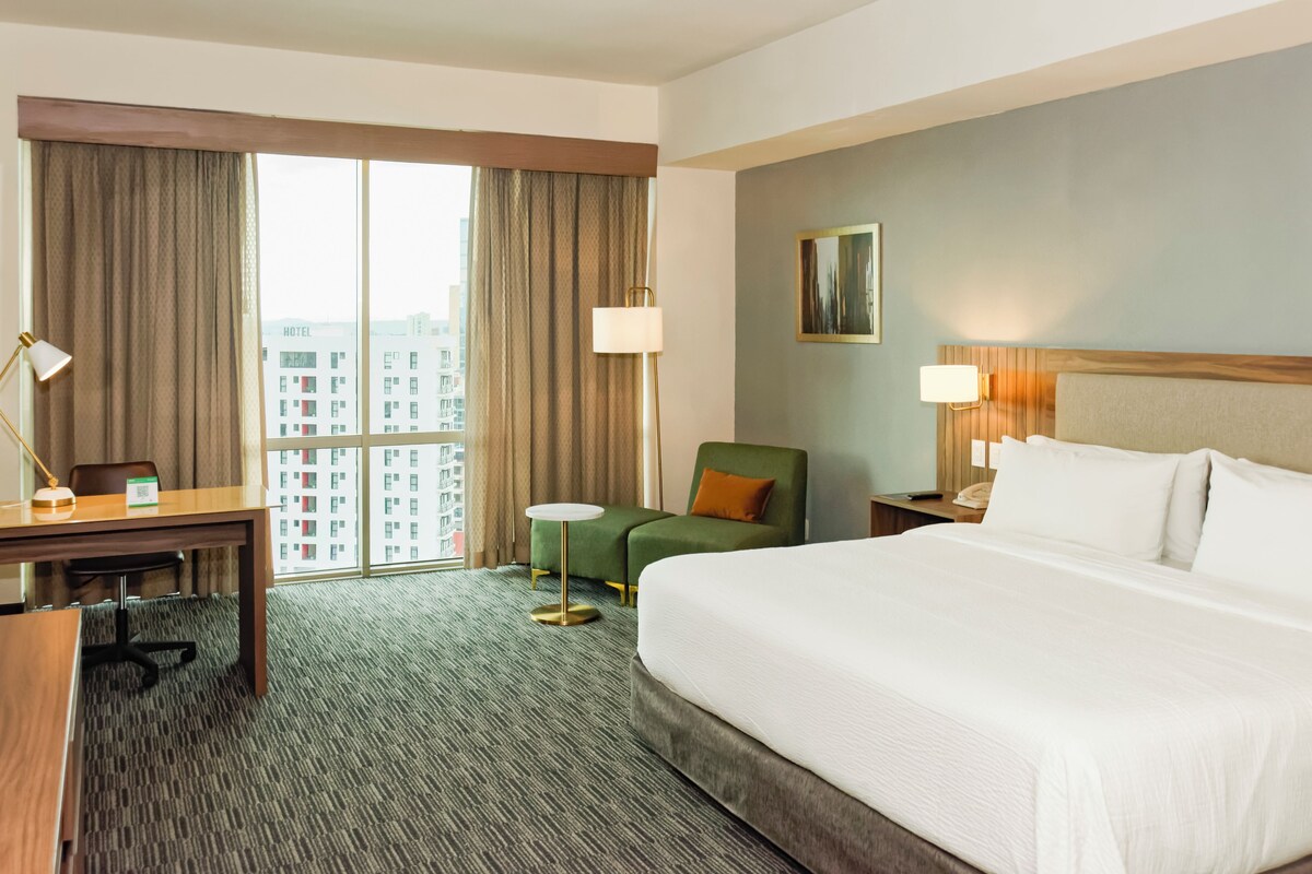 A hotel room with a large bed, a desk, a chair, a small table, and a view of the cityscape through the window.