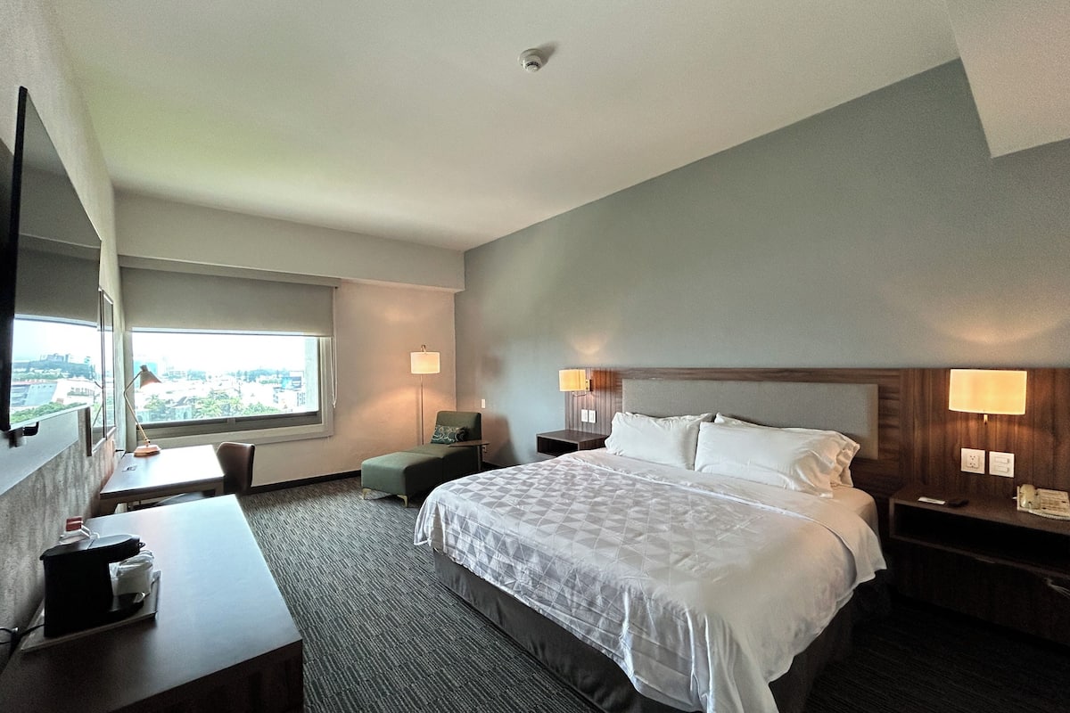 A hotel room with a bed, desk, chair, and a view of the city through a large window.