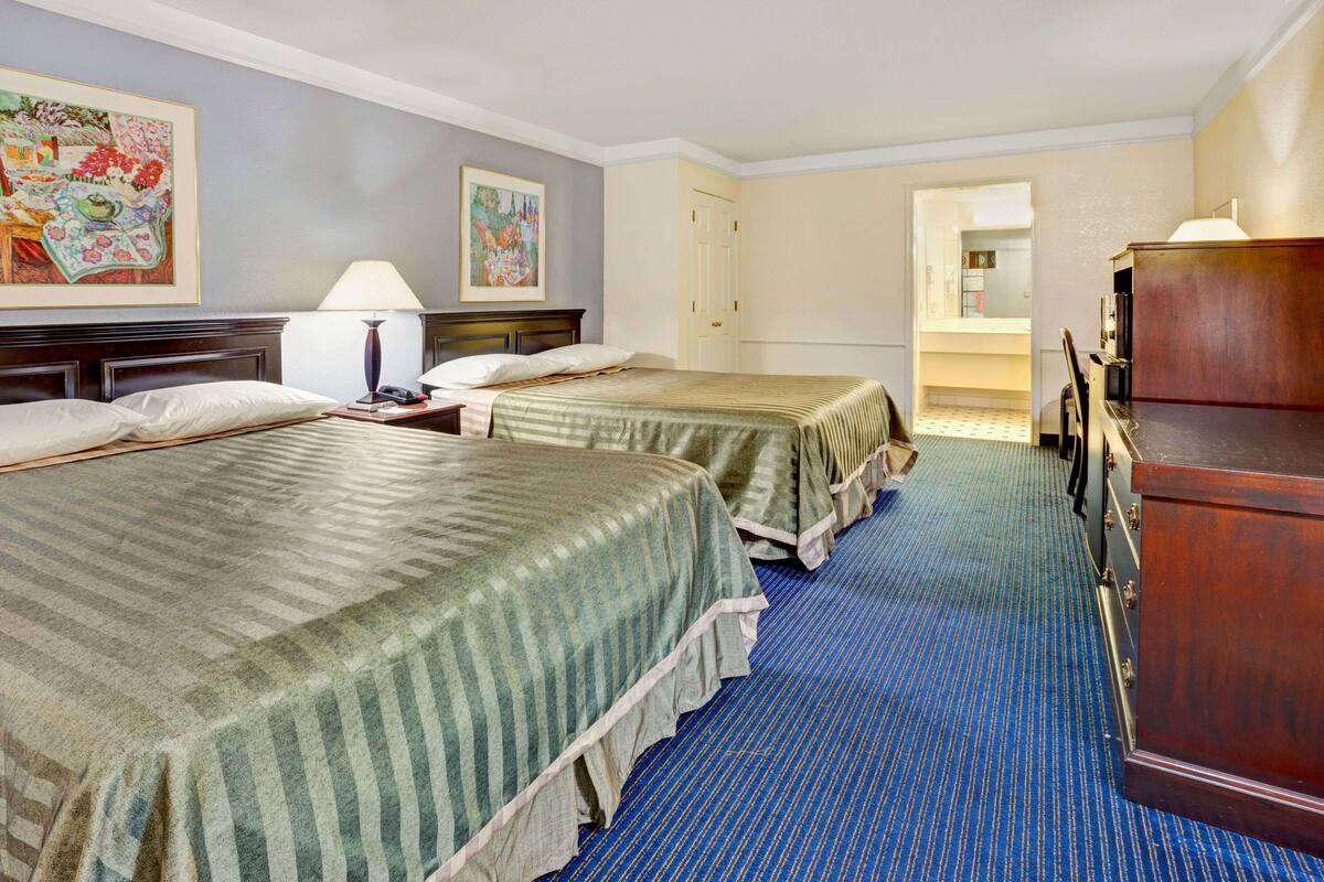 Quarto de hotel com duas camas, uma cômoda, uma escrivaninha e um abajur.