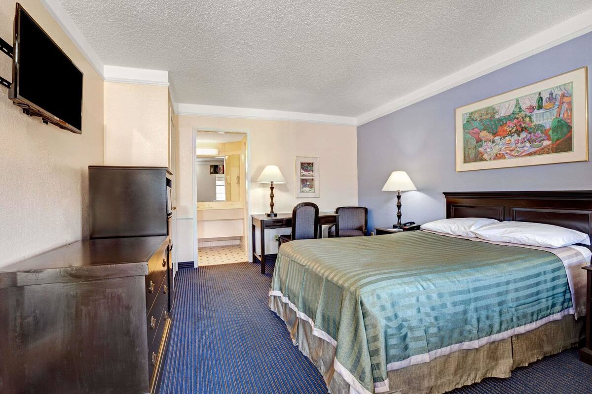 Quarto de hotel com cama, escrivaninha, cadeira, televisão e obra de arte na parede.