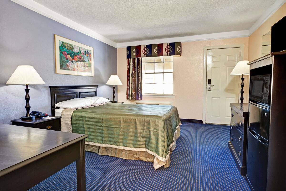 Quarto de hotel com uma cama, duas mesas de cabeceira com luminárias, um penteadeira, uma televisão, uma janela com cortinas e uma porta.