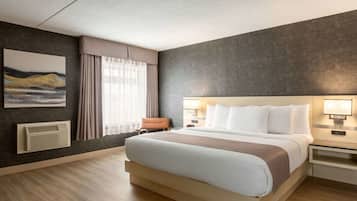 A hotel room with a large bed, a bedside table, a wall-mounted air conditioner, a window with curtains, and a painting on the wall.
