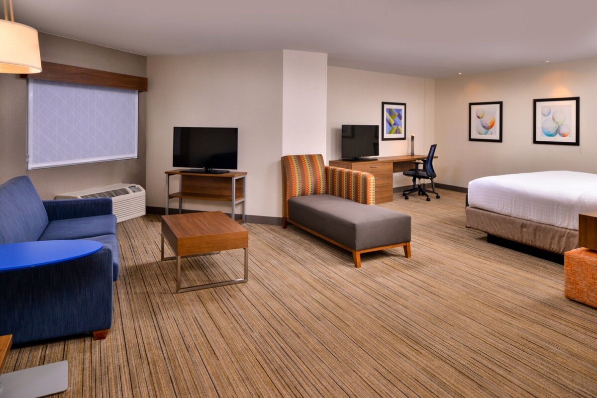 A hotel room with a bed, a sofa, a desk with a computer, a television, and a window with blinds.