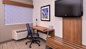 A hotel room with a wooden desk, a black office chair, a mounted flat-screen TV, a framed abstract art piece, a window with a geometric patterned blind, and a wall-mounted air conditioner. - Holiday Inn Express Walnut Creek by IHG (Walnut Creek)