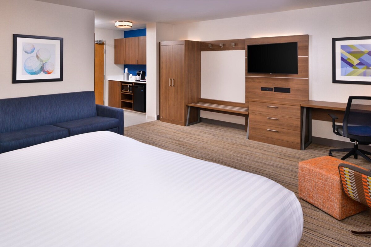 A hotel room with a large bed, a sofa, a desk with a chair, a TV, and a kitchenette area.