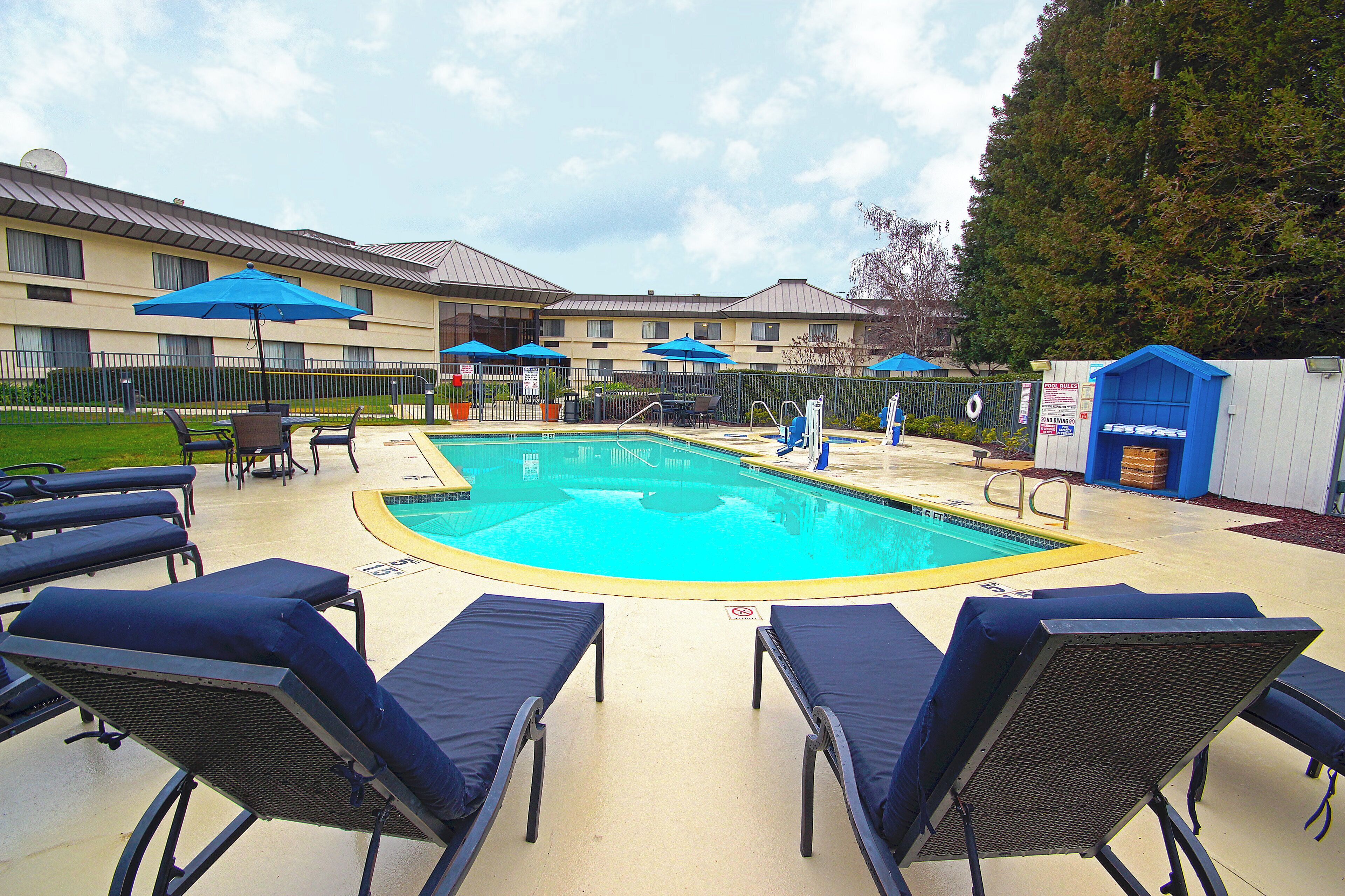 A swimming pool with lounge chairs and umbrellas, surrounded by a fence and a building with multiple windows.