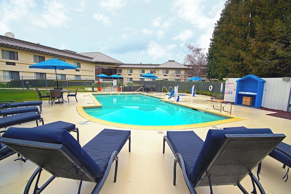 A swimming pool with lounge chairs and umbrellas, surrounded by a fence and a building with multiple windows.