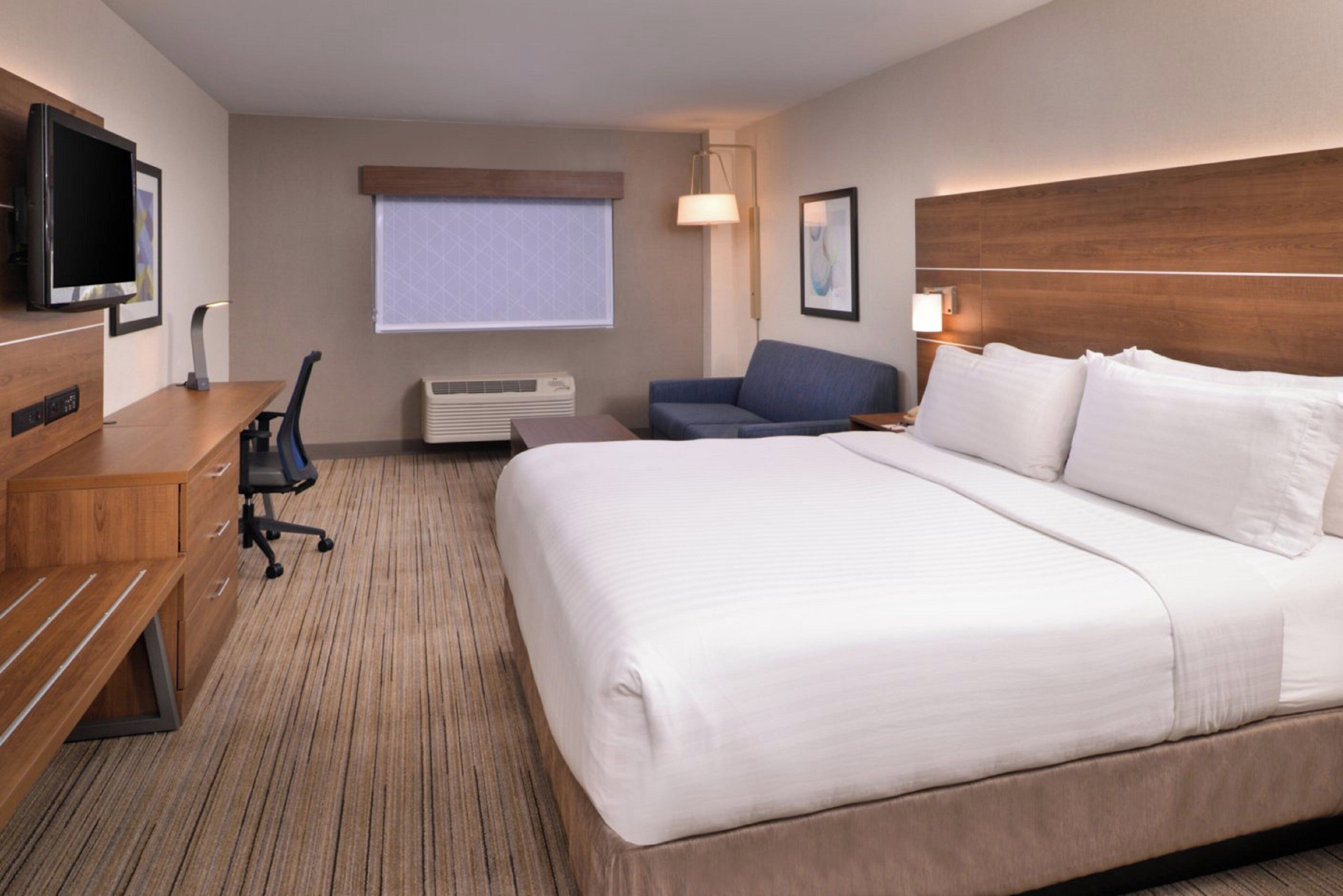 A hotel room with a bed, a desk, a chair, a TV, an air conditioning unit, and a window with blinds.