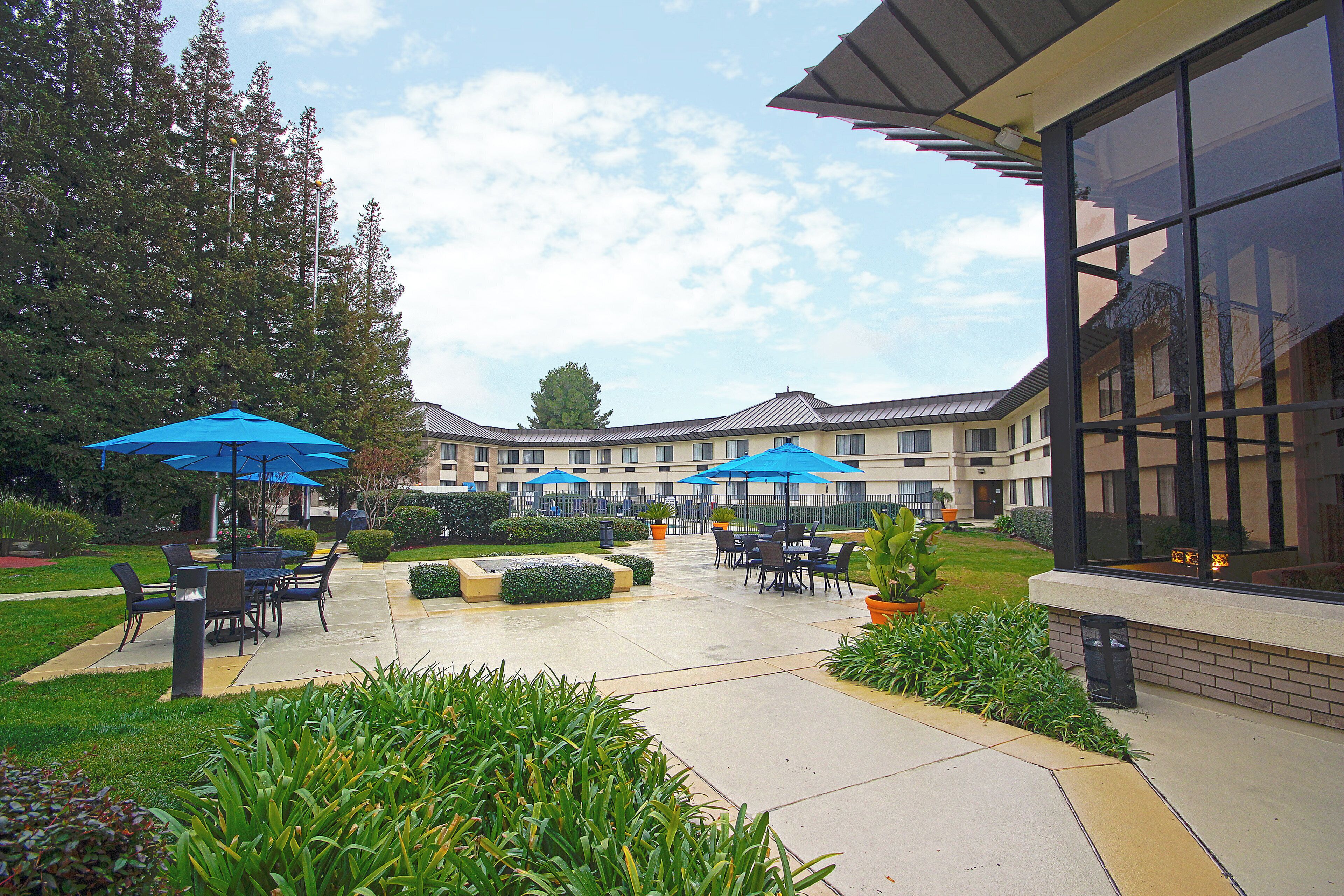 An outdoor patio area with seating and umbrellas, surrounded by greenery and a building with large windows.