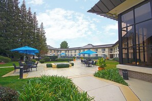 An outdoor patio area with seating and umbrellas, surrounded by greenery and a building with large windows. - Holiday Inn Express Walnut Creek by IHG (Walnut Creek)