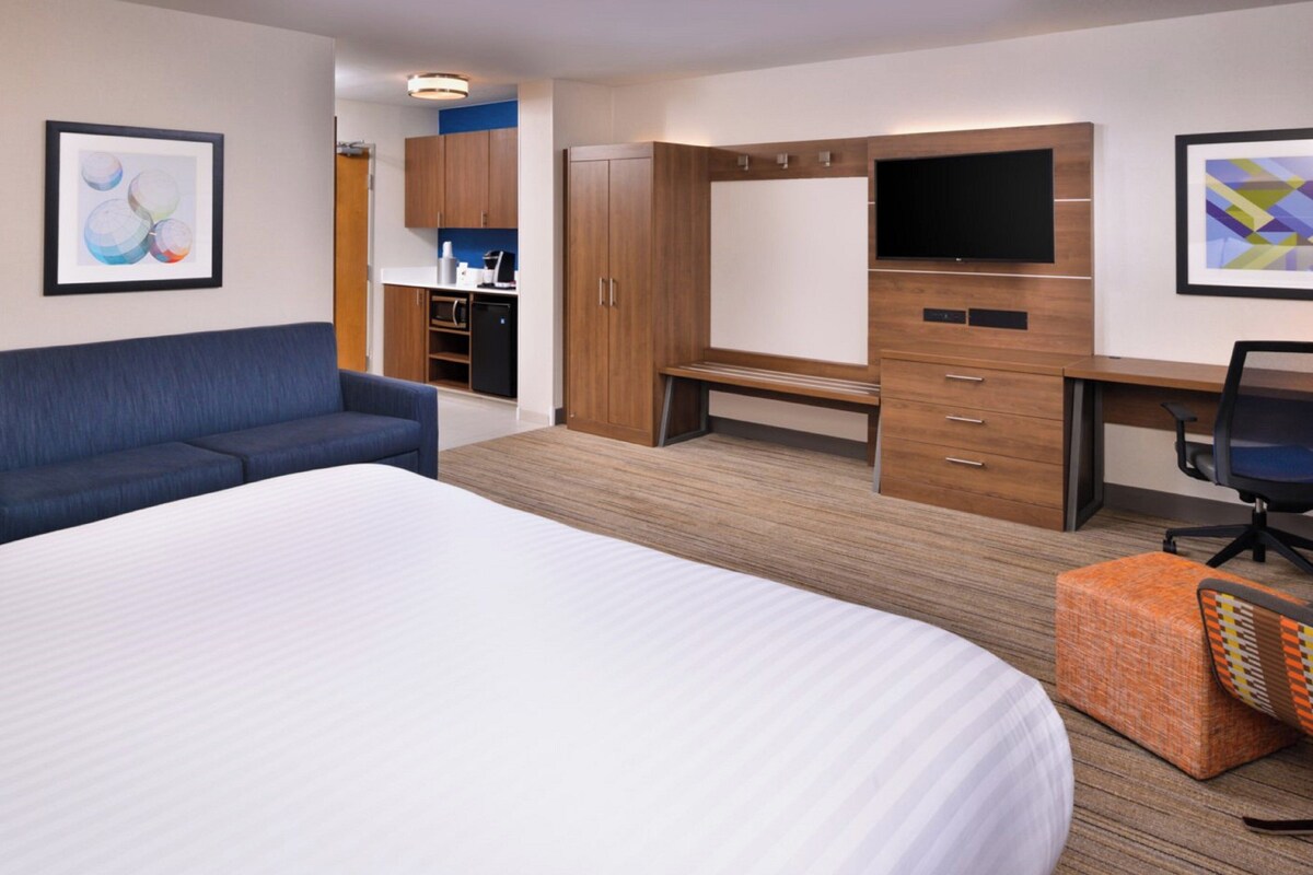 A hotel room with a large bed, a sofa, a desk with a chair, a TV, and a kitchenette.