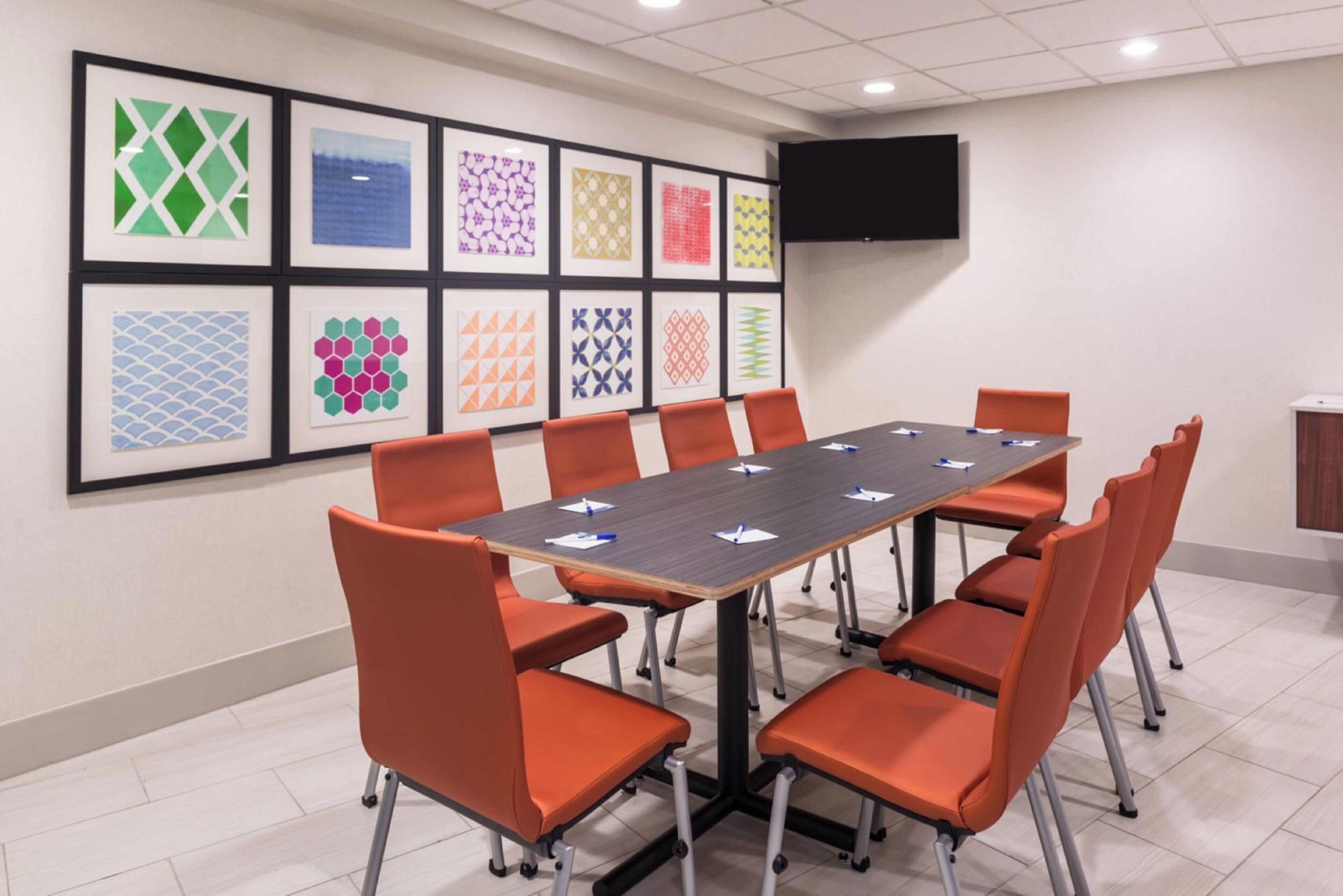 A conference room with a long table and orange chairs, a tiled floor, and a wall with framed geometric patterns.