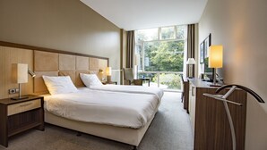 Une chambre d’hôtel avec un grand lit, des tables de chevet avec des lampes, un bureau avec une chaise et une vue sur la verdure par la fenêtre.