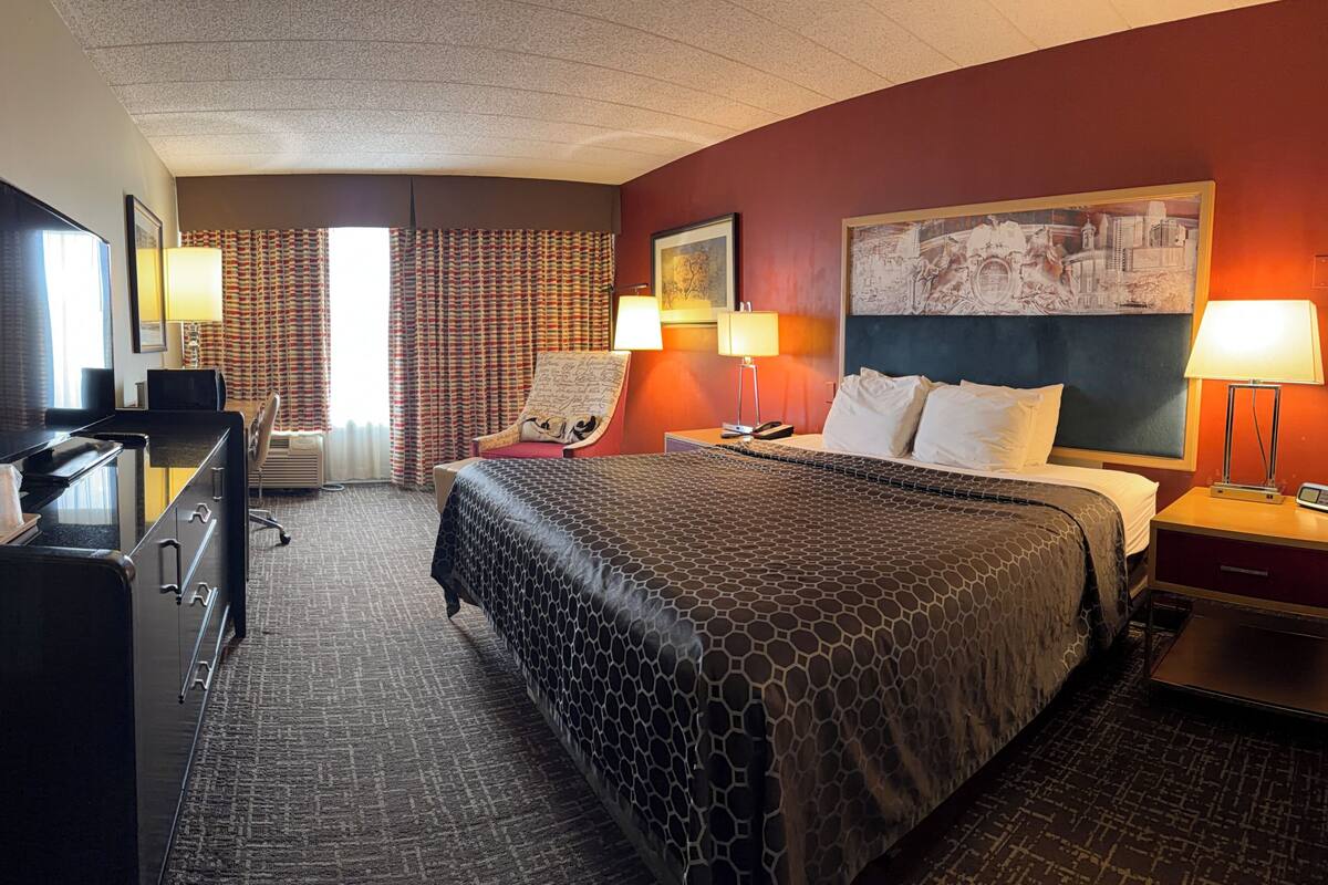 A hotel room with a bed, desk, chair, and a painting on the wall.