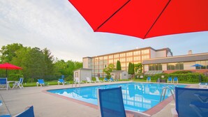 Une piscine entourée de chaises longues et de parasols, elle-même bordée d’un bâtiment aux grandes fenêtres et d’une zone délimitée par une clôture, où abondent les plantes et la verdure.