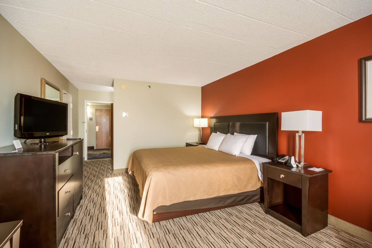 A hotel room with a bed, a television on a dresser, a nightstand with a lamp, and a door leading to another room.
