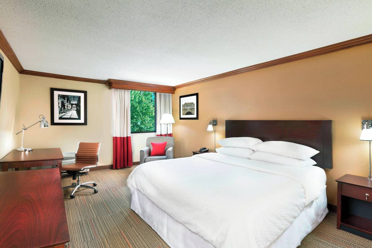 Une chambre d’hôtel avec un grand lit, un bureau, une chaise, une table de chevet et une fenêtre avec des rideaux.