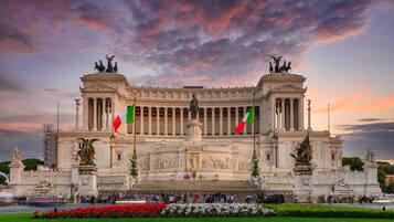 A grand neoclassical building with columns and statues, surrounded by a well-maintained garden and flags.