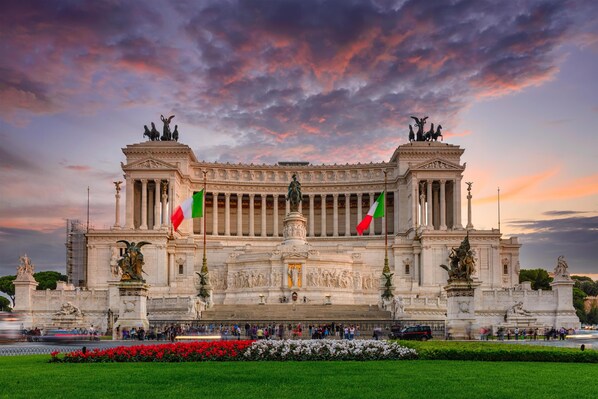 A grand neoclassical building with columns and statues, surrounded by a well-maintained garden and flags.