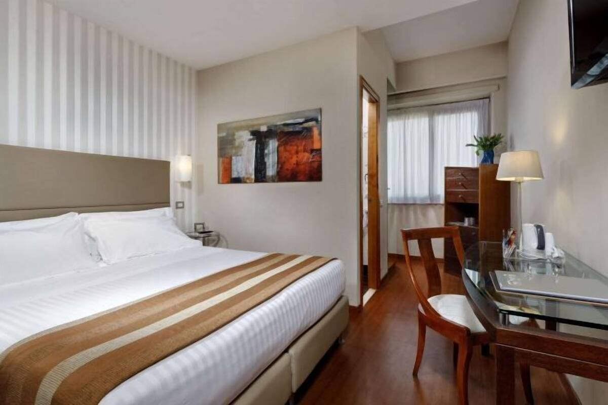 A hotel room with a bed, a desk, a chair, a lamp, and a painting on the wall.