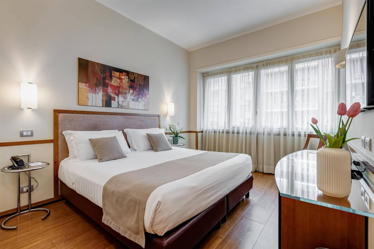 A neatly arranged hotel room with a large bed, bedside tables, a desk, and a window with curtains.
