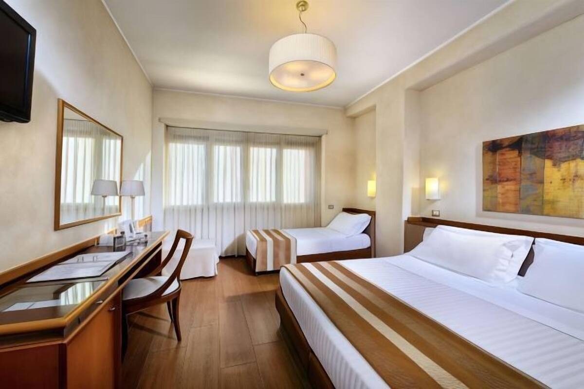 A hotel room with a desk, two beds, a chair, a TV, and a large painting on the wall.