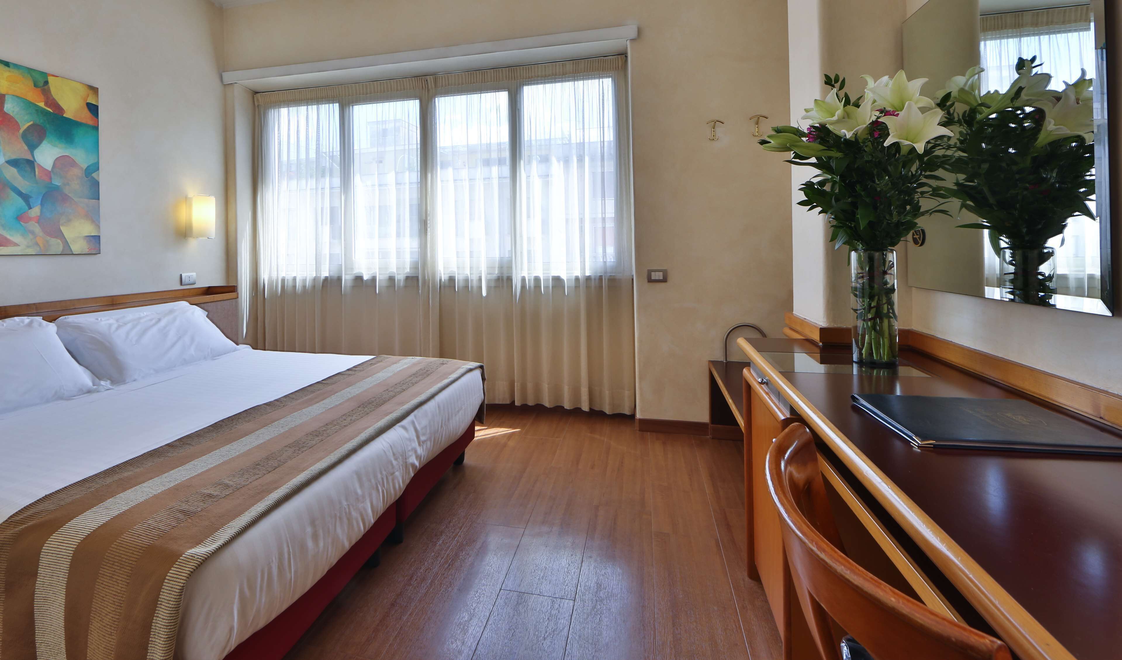 Une chambre d’hôtel avec un lit, un bureau, une chaise, une fenêtre avec des rideaux, un vase de fleurs et un tableau au mur.
