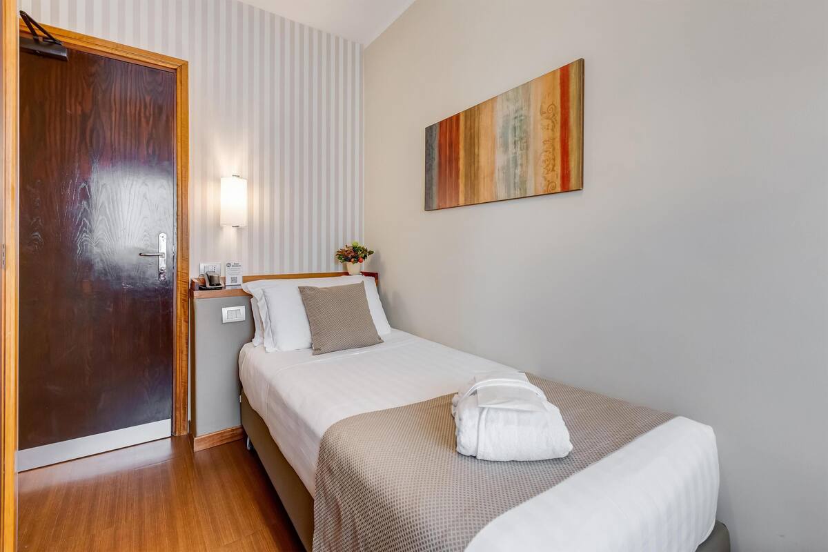 A neatly arranged hotel room with a wooden door, a bed with a white and beige bedspread, a wall-mounted painting, and a bedside table with a lamp and a small plant.