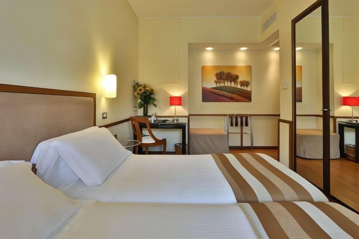 A hotel room with two beds, a desk, a chair, a mirror, and a painting on the wall.