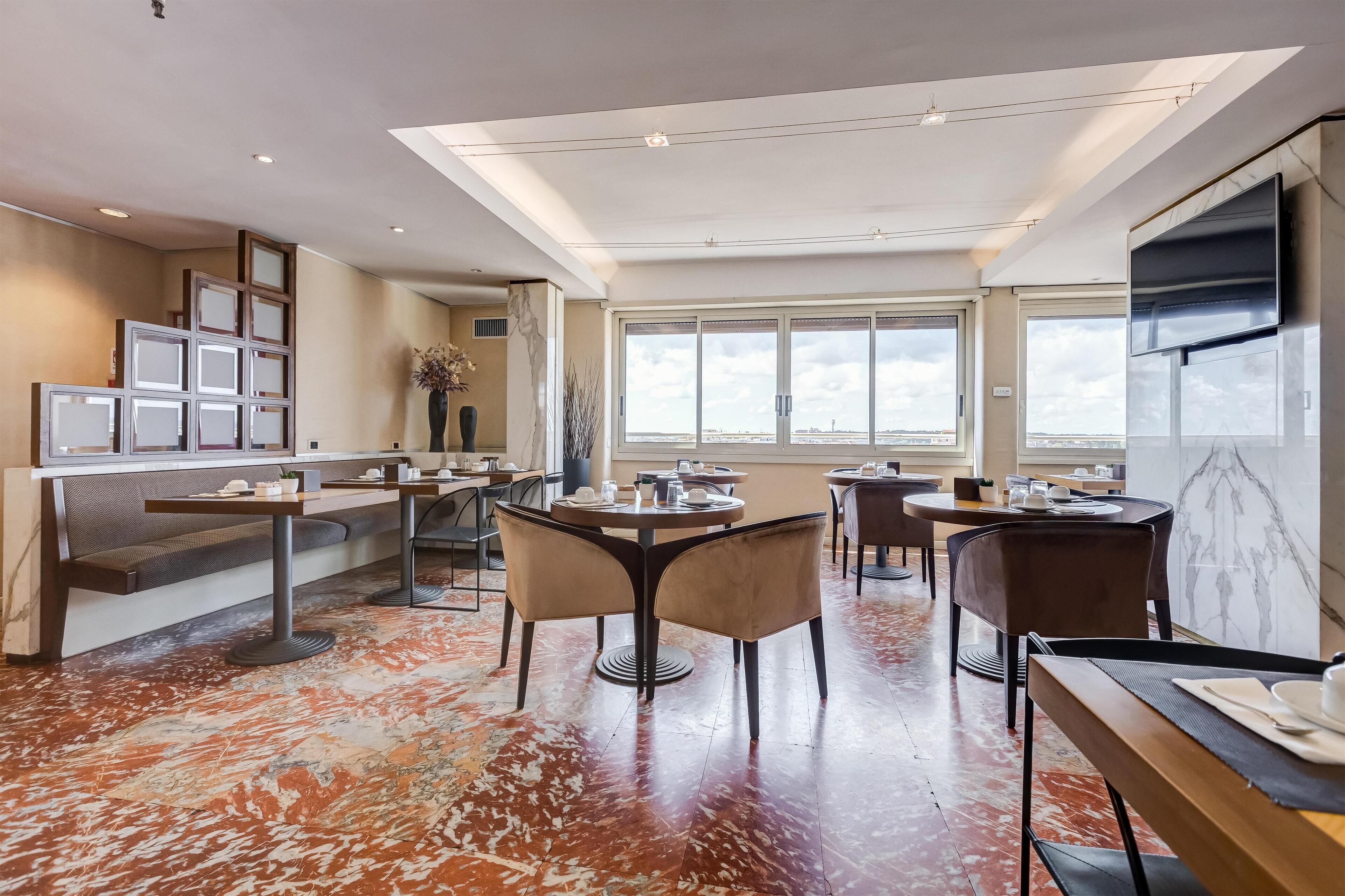 Un comedor de hotel moderno con varias mesas y sillas, un ventanal con vistas y un techo con iluminación empotrada.