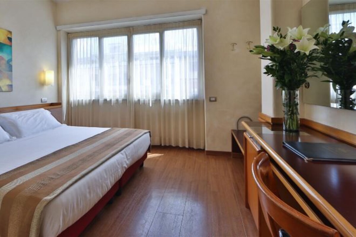 Habitación de hotel con cama, escritorio, silla, un jarrón con flores y ventana con cortinas.