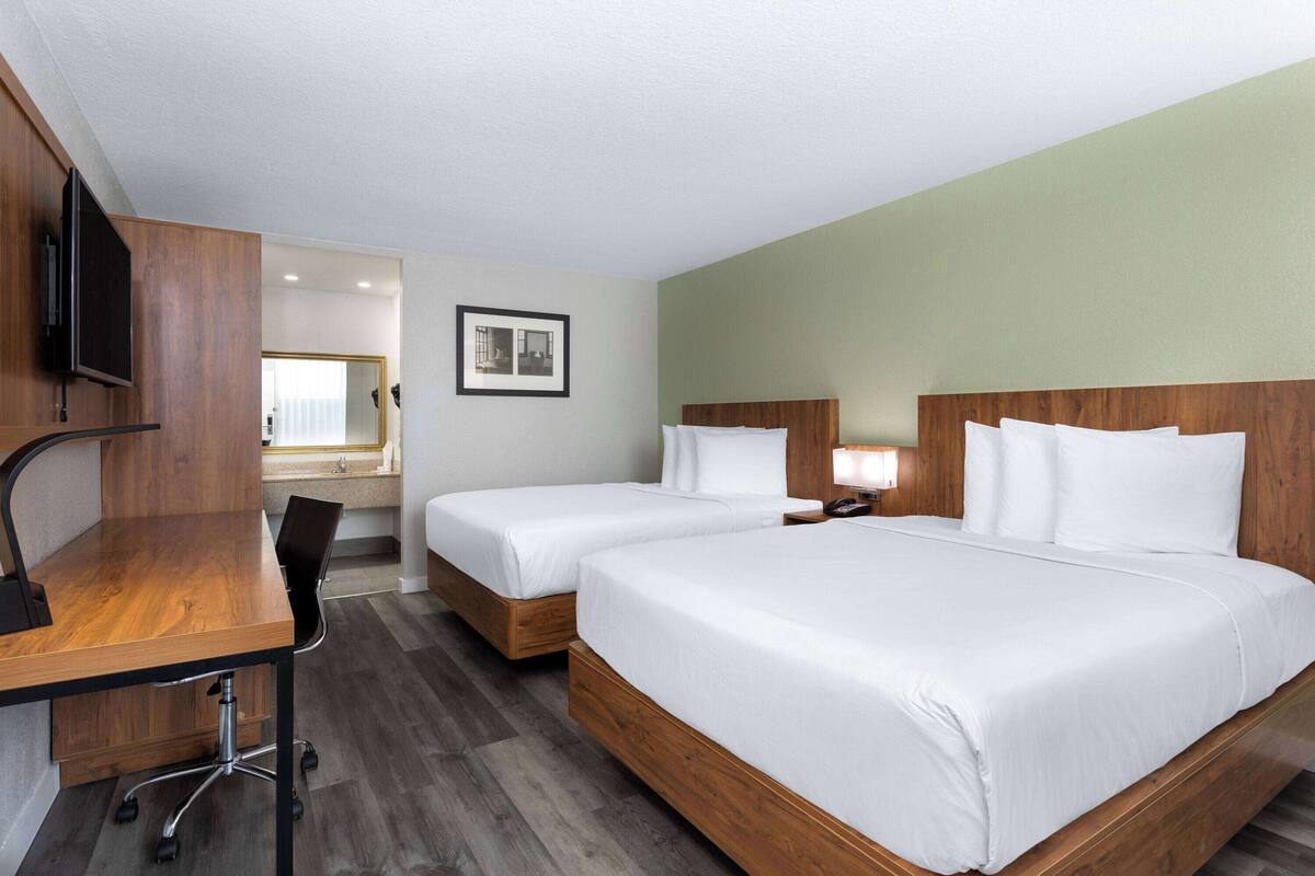 A hotel room with two single beds, a desk with a chair, a television, and a bathroom visible through an open door.