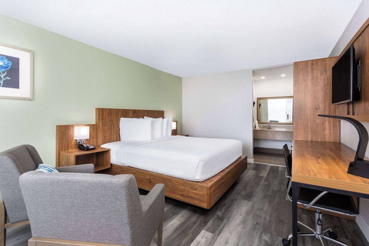 A hotel room with a wooden bed, a desk with a chair, a TV, and a bathroom visible in the background.