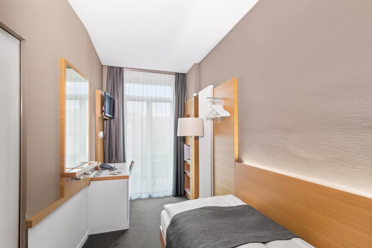 A hotel room with a bed, a desk with a chair, a television, a mirror, a window with curtains, and a wardrobe.