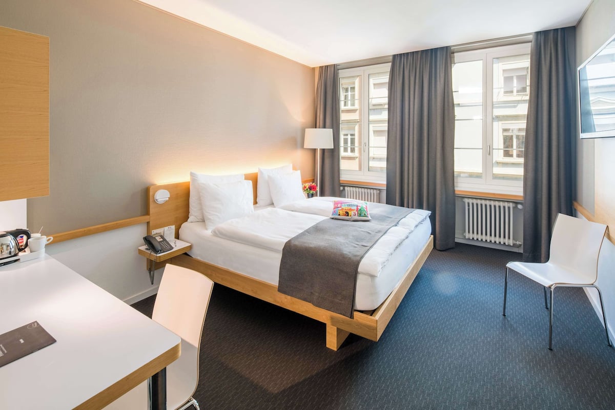 A hotel room with a bed, bedside table, lamp, chair, and a view of the city through the window.