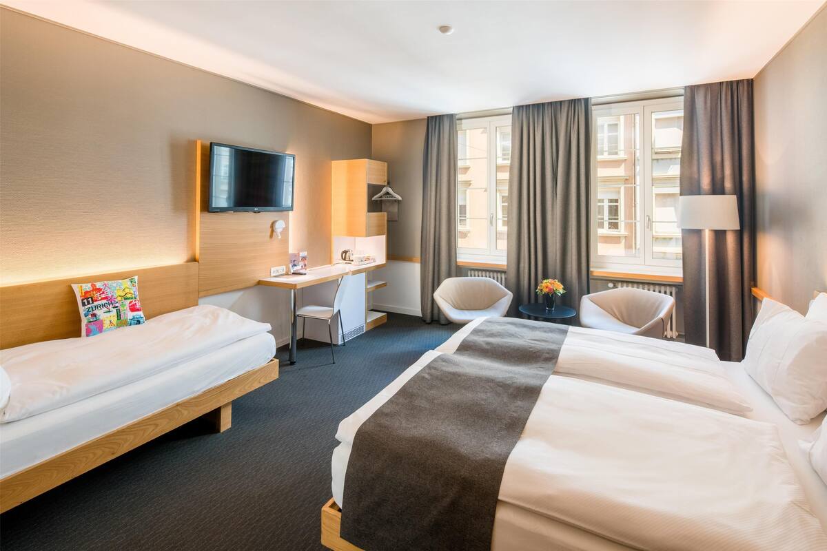 A hotel room with two beds, a desk, a chair, a TV, and a window with curtains.