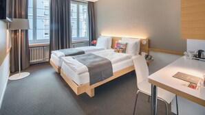 Une chambre d’hôtel avec deux lits, un bureau, une chaise, une lampe, un téléviseur, une fenêtre avec des rideaux et un radiateur.