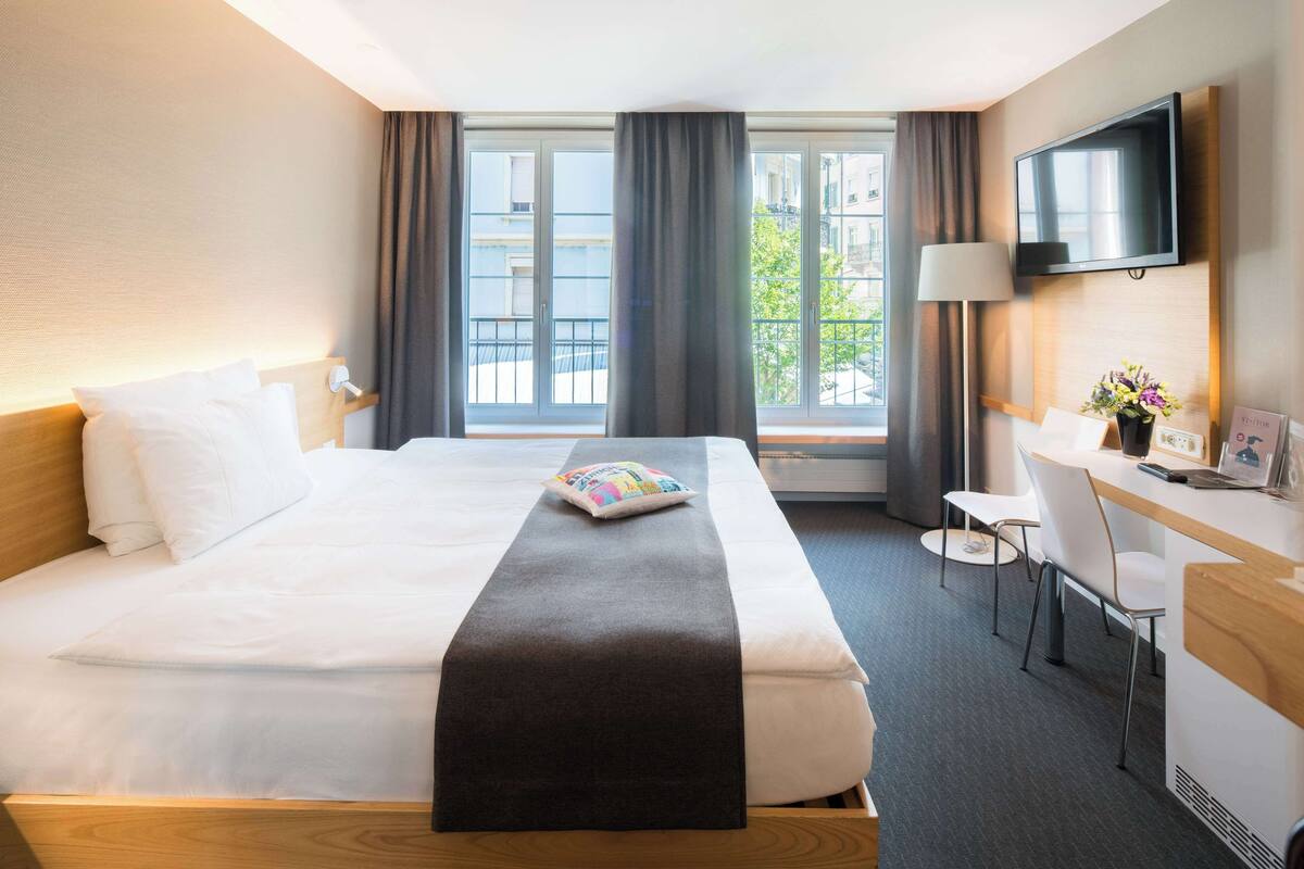 A hotel room with a bed, a desk, a chair, a TV, and a window with curtains.
