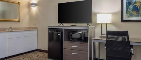 Habitación de hotel con kitchenette, un televisor de pantalla plana, un escritorio con silla, una lámpara y un cuadro en la pared.