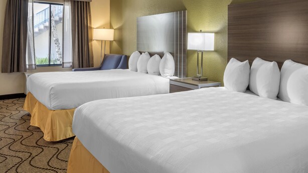 A hotel room with two beds, a desk, a chair, a lamp, and a view of a building outside. - Best Western Heritage Inn (Concord)
