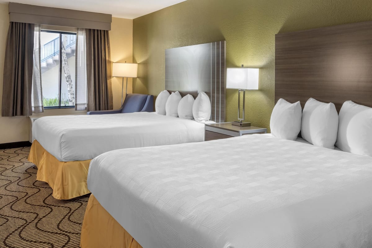 A hotel room with two beds, a desk, a chair, a lamp, and a view of a building outside.