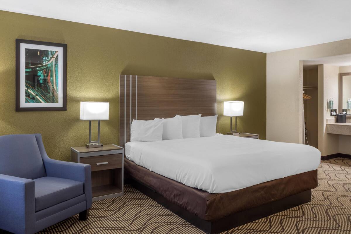 A hotel room with a large bed, two bedside tables with lamps, a chair, a framed picture on the wall, and a patterned carpet.