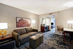 Habitación de hotel con sofá, reposapiés, una mesa de comedor con sillas, zona de cocina y un cuadro grande en la pared.
