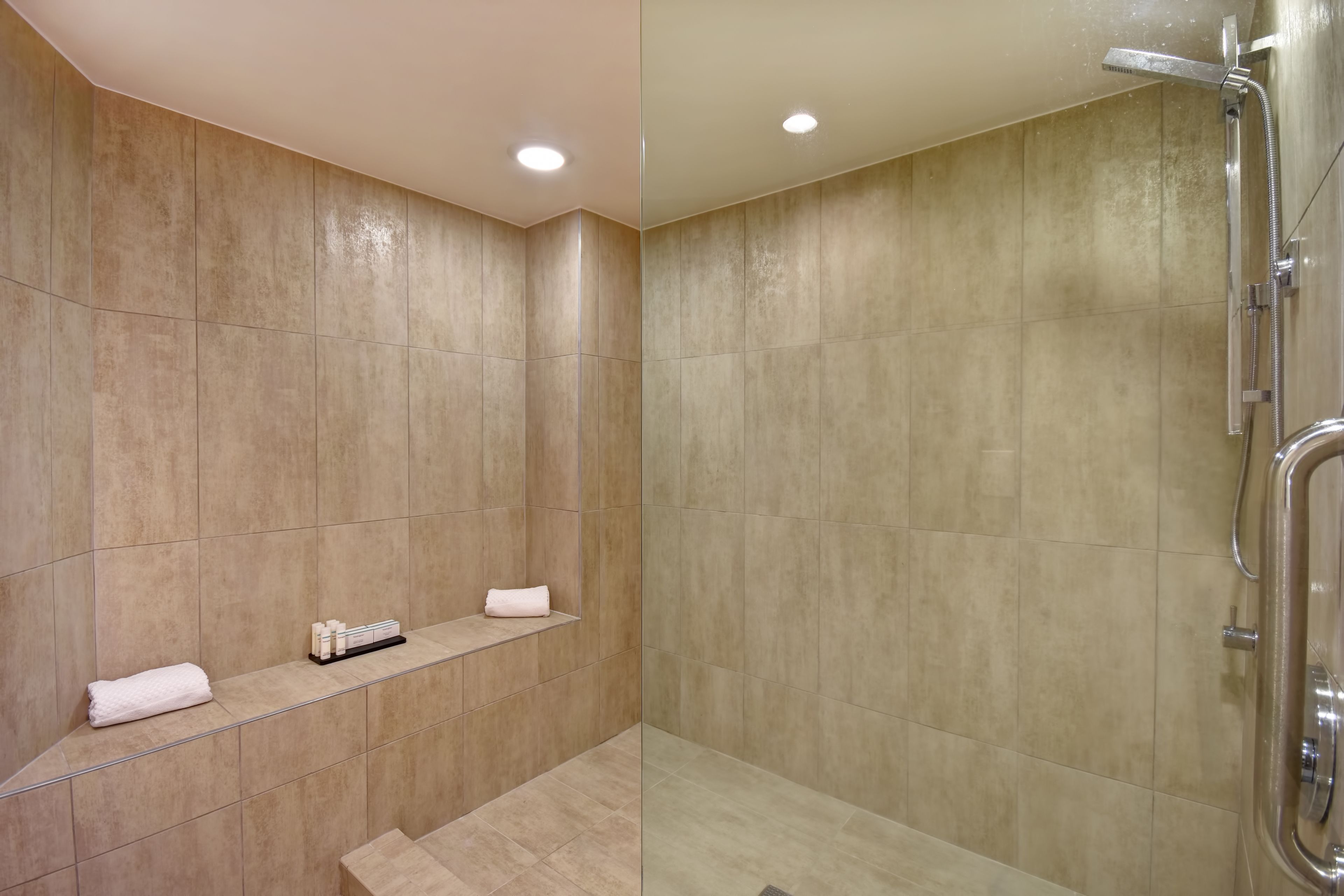 A modern bathroom with beige tiles, a glass shower enclosure, and a shelf with toiletries.