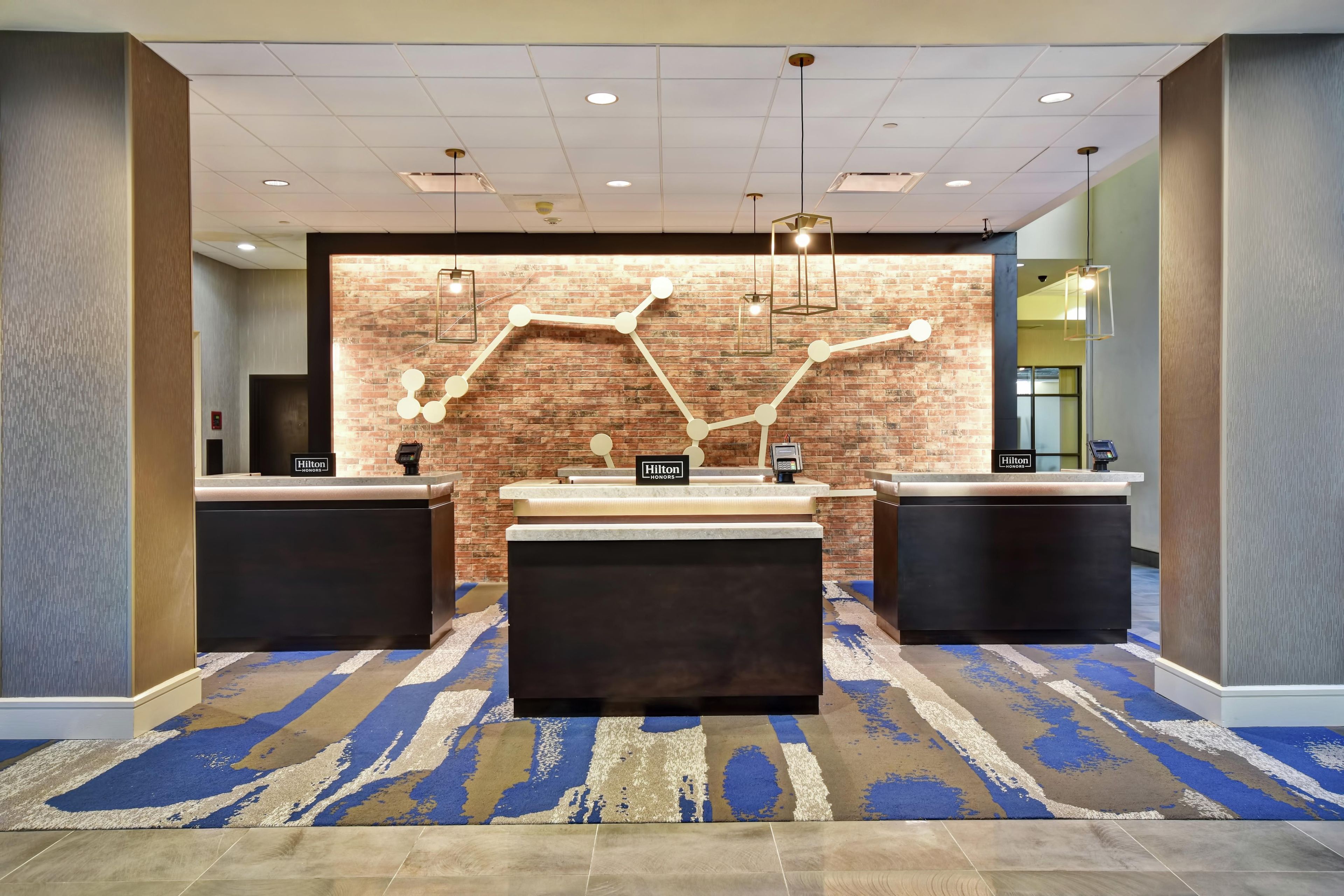 A hotel reception area with a brick wall, two reception desks, a central seating area, and a patterned carpet.