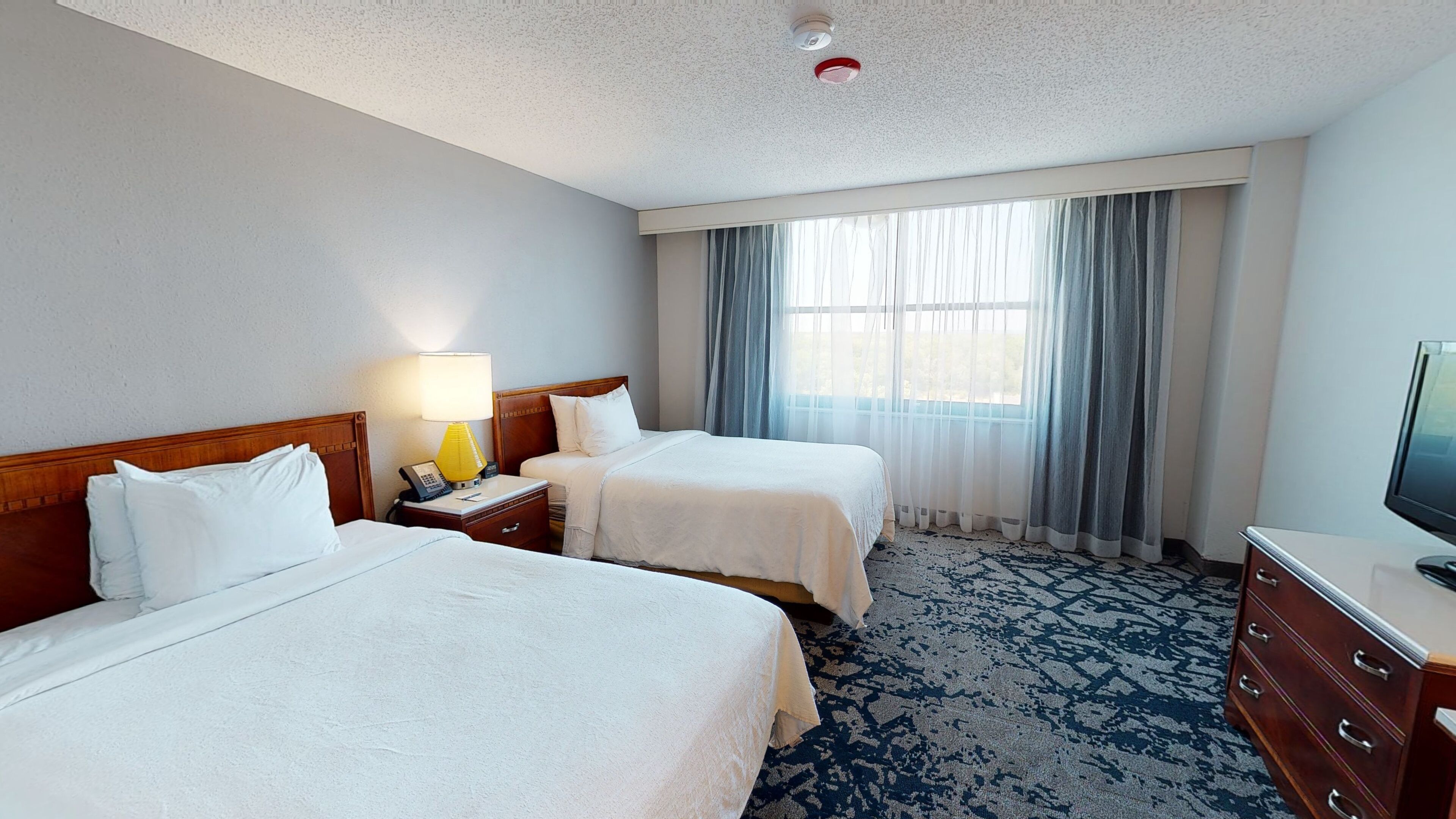 Quarto de hotel com duas camas, uma escrivaninha com luminária, televisão e uma janela grande com cortinas.