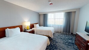 Habitación de hotel con dos camas, un escritorio con lámpara, televisión y un amplio ventanal con cortinas.