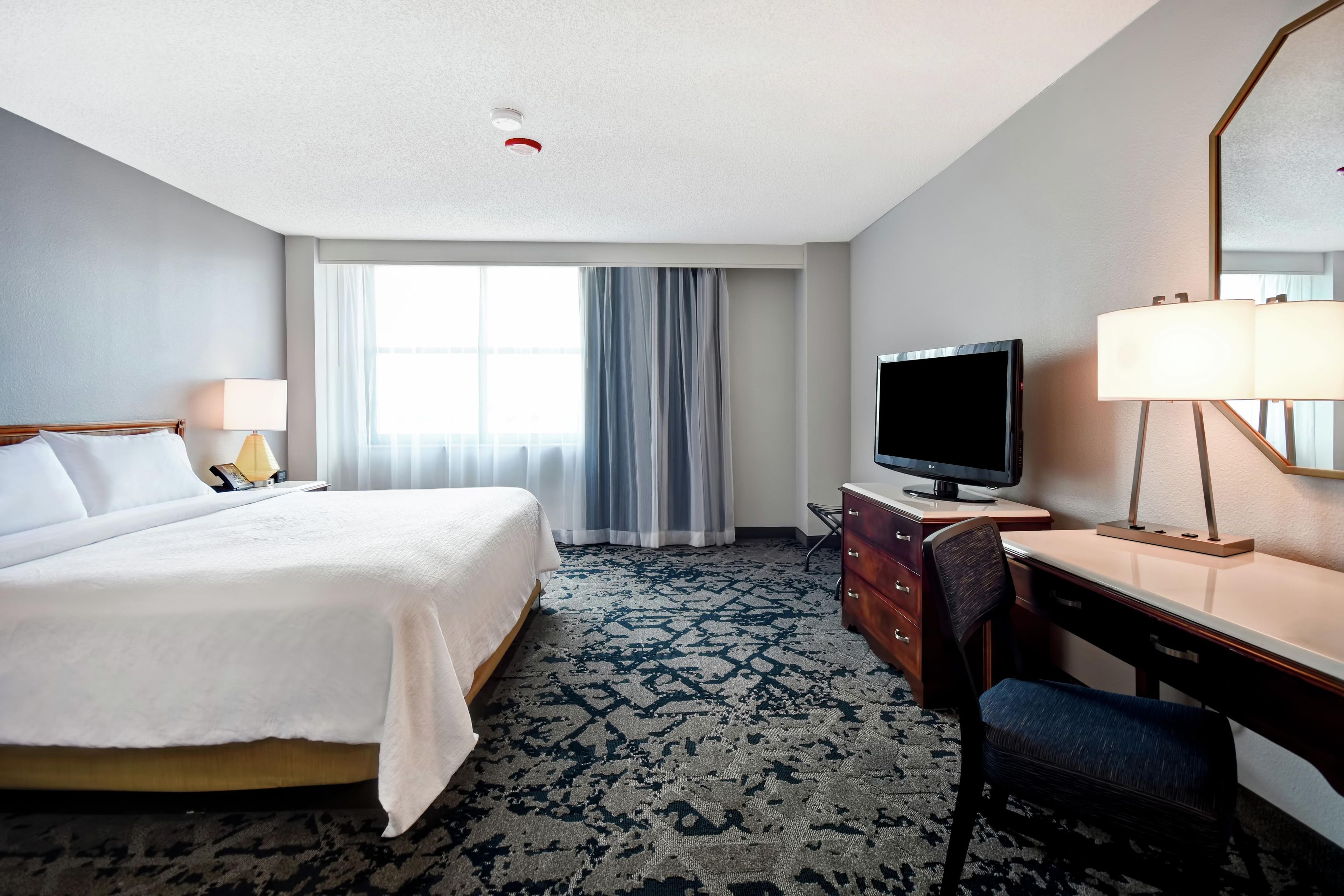 A hotel room with a bed, a desk with a TV, a chair, a lamp, and a window with curtains.